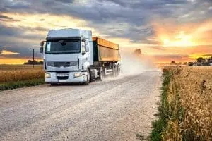 A white semi-truck drives on a gravel road through a rural landscape at sunset, with dust trailing behind and fields on either side.