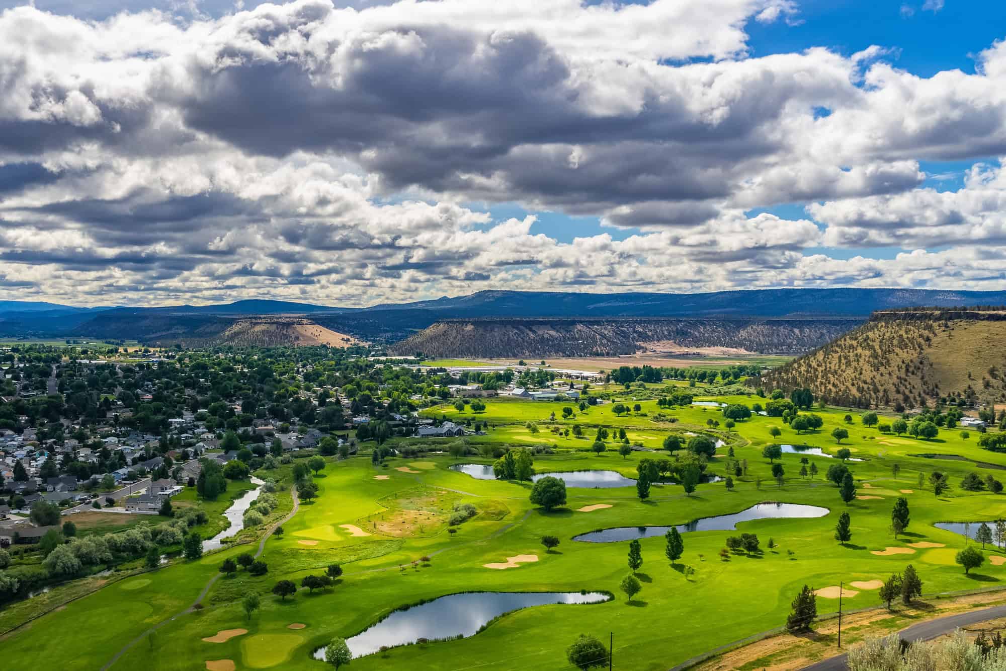 Aerial view of a golf course with lush green fairways, several ponds, and scattered trees. Adjacent residential area and hills in the background under a partly cloudy sky.