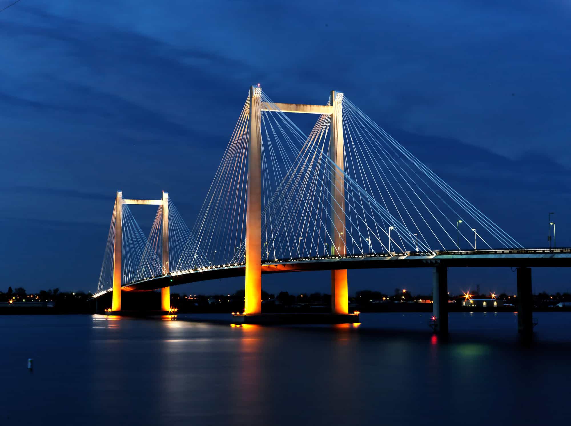 A cable-stayed bridge illuminated at night, spanning across a body of water with a deep blue sky in the background.
