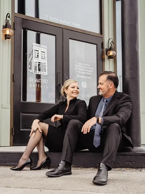 Two people in business attire sit on steps outside DS Watkins Gallery. The woman smiles at the man, and both wear black suits. The gallery entrance features glass doors and lanterns.