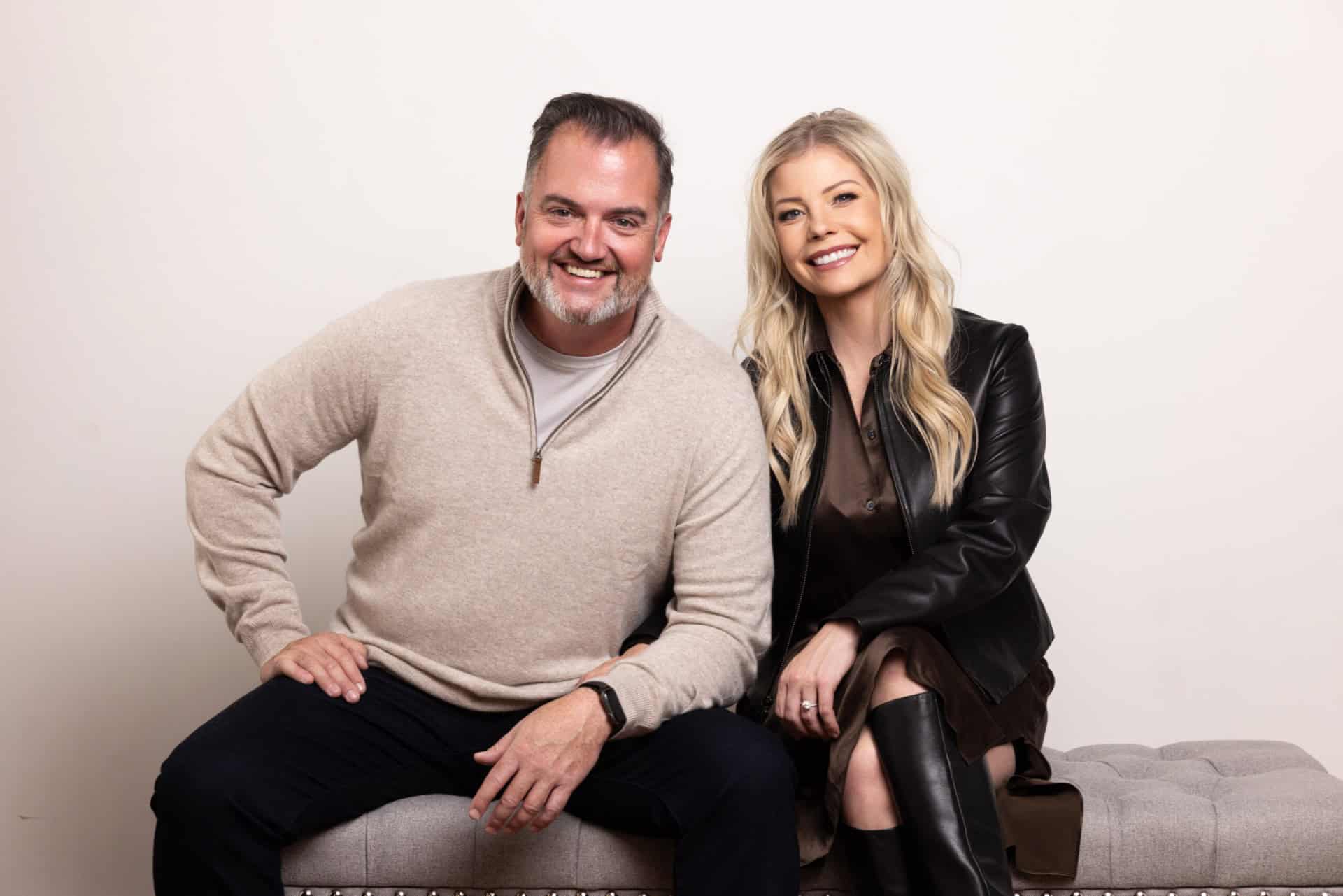 A man and woman are seated on a bench. The man wears a beige sweater, and the woman wears a black leather jacket. Both are smiling at the camera against a plain white background.