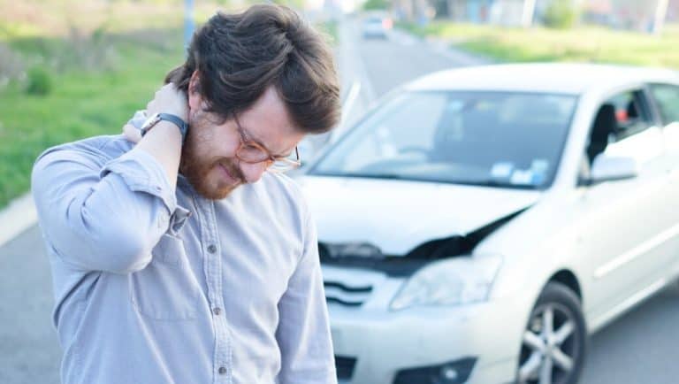 A man holds his neck in discomfort near a damaged car on the roadside.