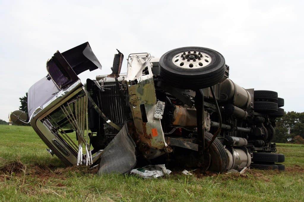 Overturned semi-truck lying on its side in a grassy area, showing damaged front and exposed undercarriage.
