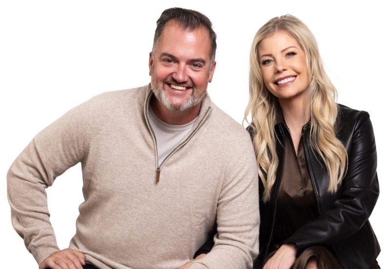 A man in a beige sweater and a woman in a black leather jacket sit side by side, both smiling at the camera against a white background.