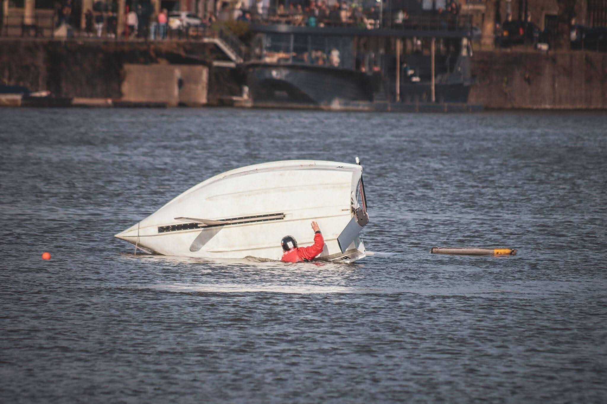 A capsized white boat in the water with a person in a red jacket holding onto the hull; onlookers gather on a riverside embankment in the background