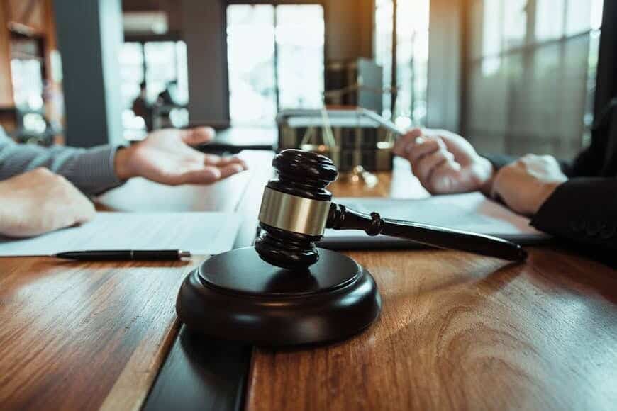 A gavel rests on a wooden table. In the background, a lawyer sits across the table from their client.