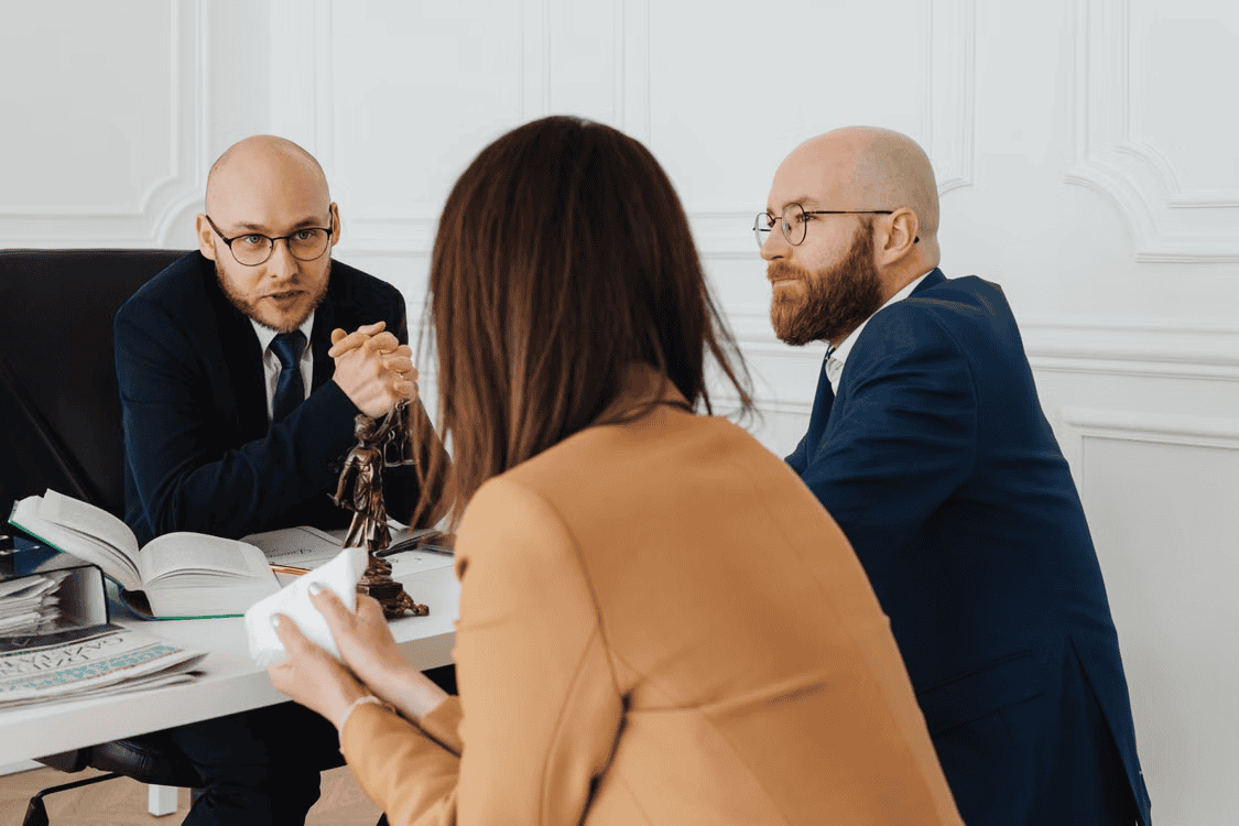 A man wearing glasses and a business suit talks to a couple across from a desk.