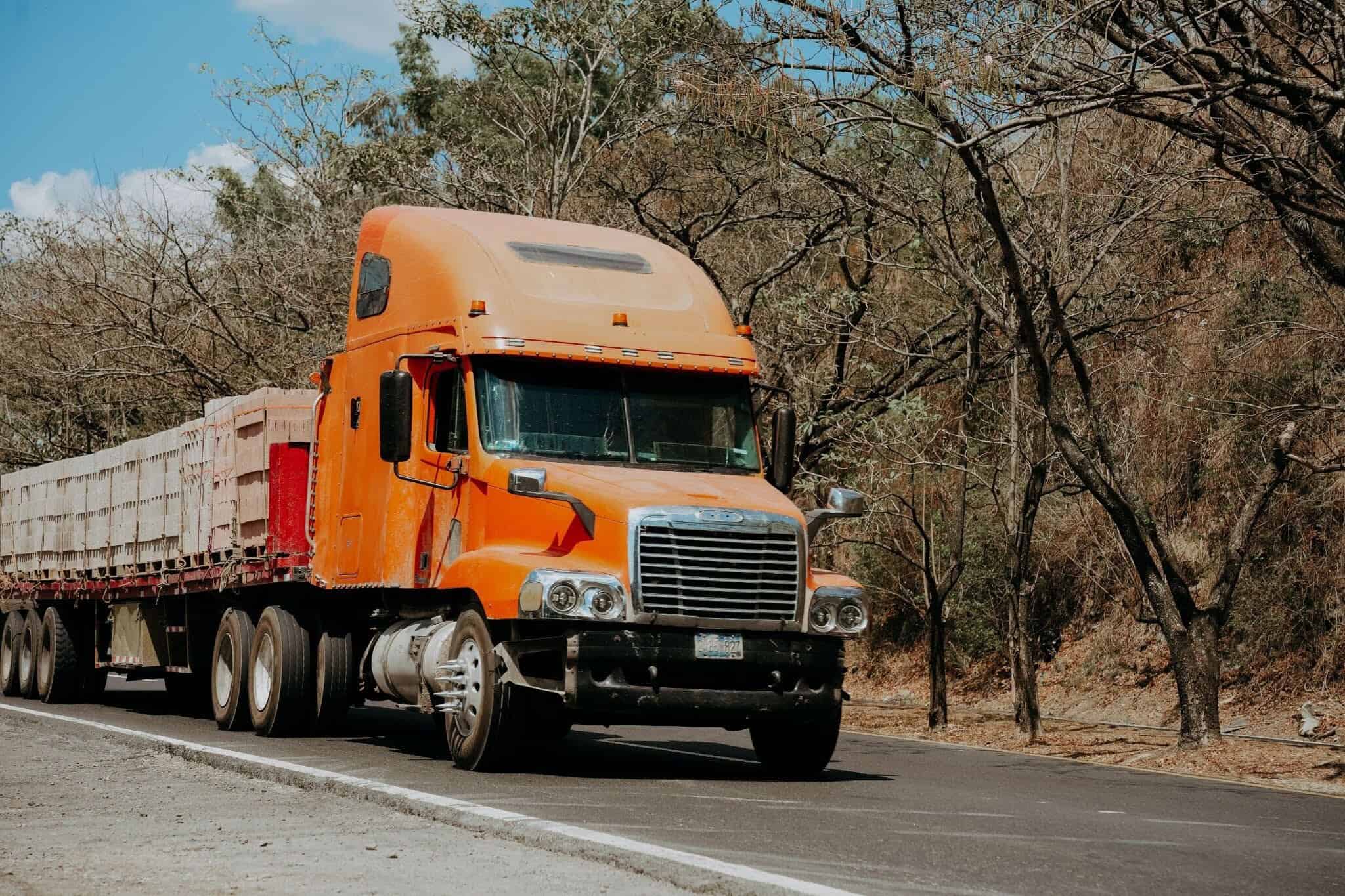A heavy-duty orange Freightliner semi-truck hauling a flatbed trailer travels along a rural highway.