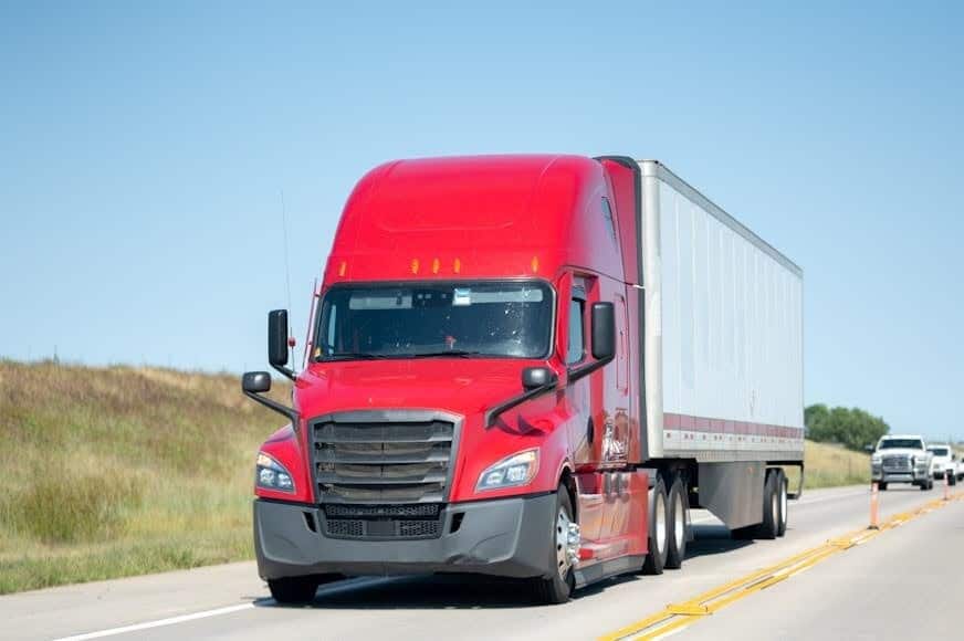 A red semi-truck is driving on the highway. A line of cars follows it.