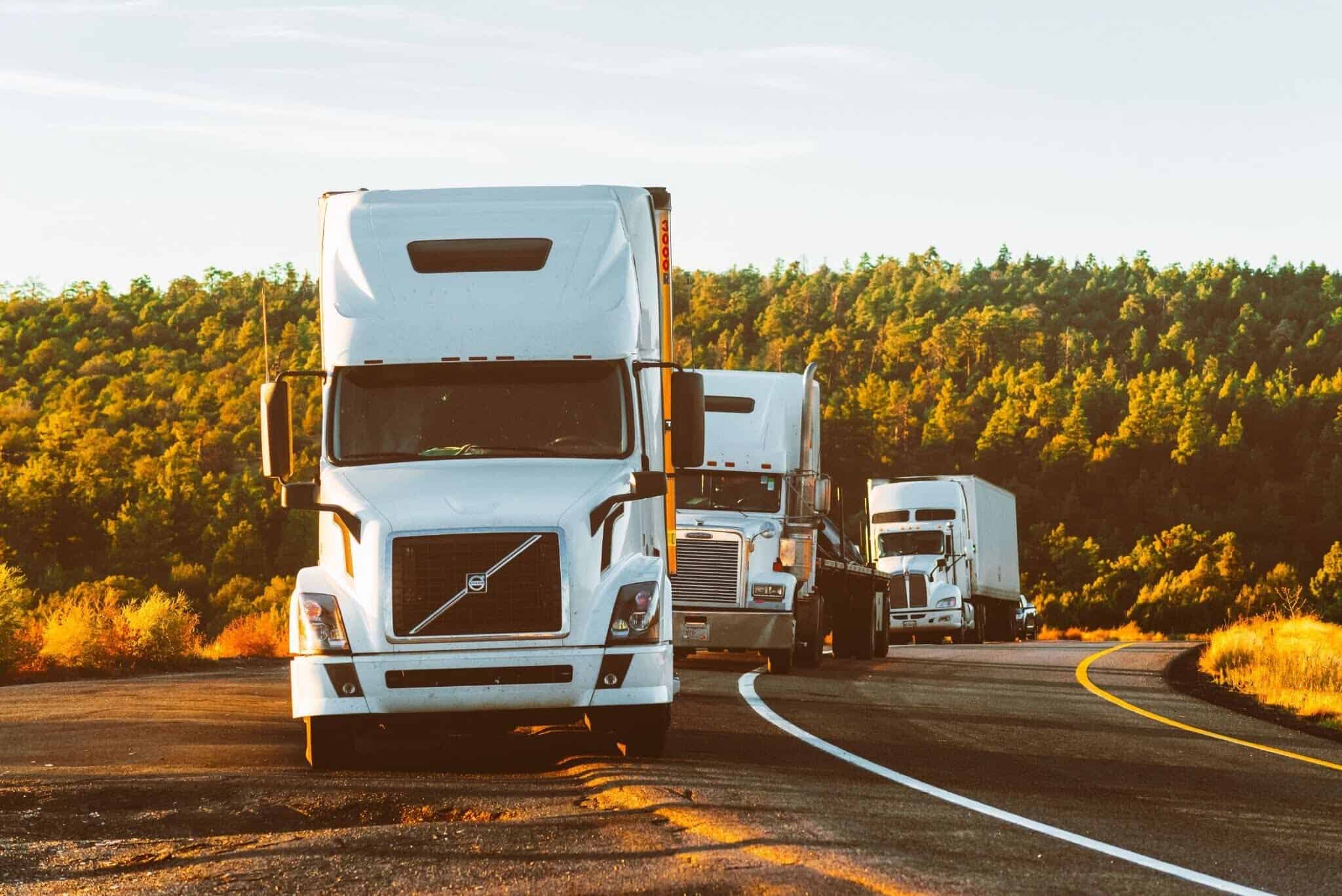 Three white semi-trucks travel in a convoy along a winding two-lane highway at golden hour.