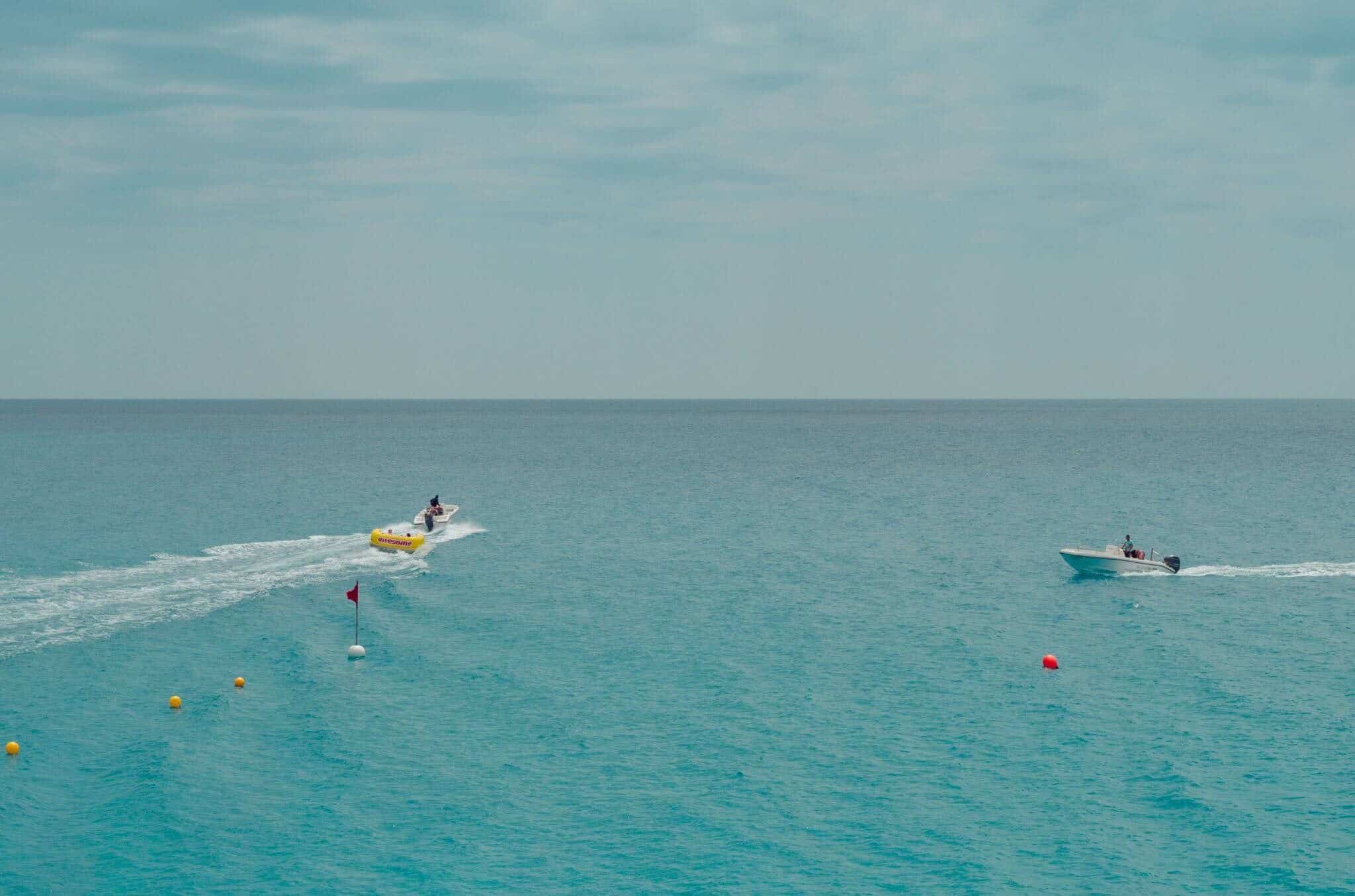 Two motorboats on a calm turquoise sea, one towing a yellow inflatable tube, with marker buoys and a red flag floating nearby under a hazy sky