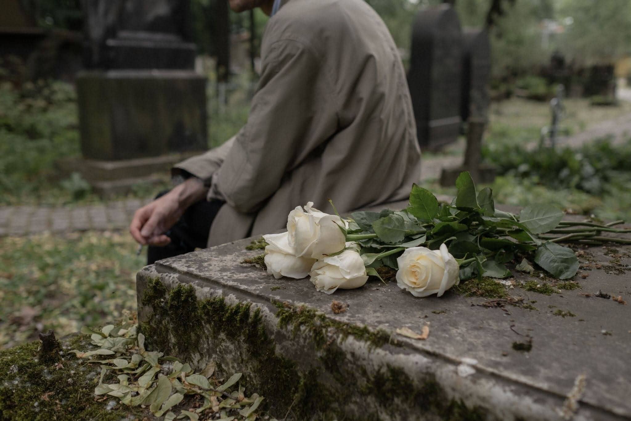 A person sits beside a moss-covered grave with white roses.