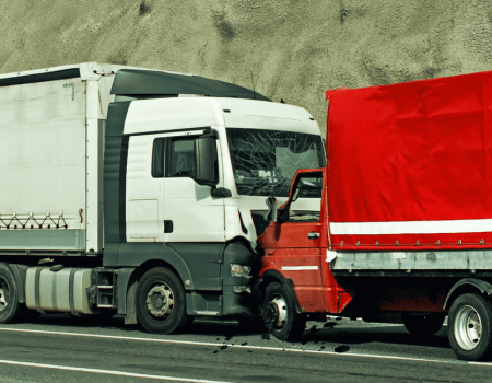 Two trucks have collided on a road. The front of a white truck is damaged and pressed against the rear of a red truck. Debris is scattered on the ground.