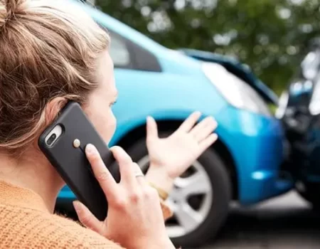 A woman uses a smartphone to call for assistance after a rear-end collision involving two cars on a road.