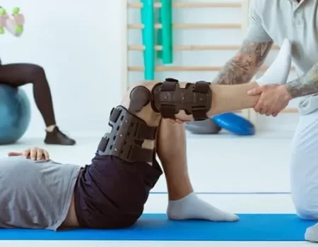 Physical therapist assists a patient with a knee brace during a rehabilitation session. Another person exercises in the background with dumbbells while seated on an exercise ball.